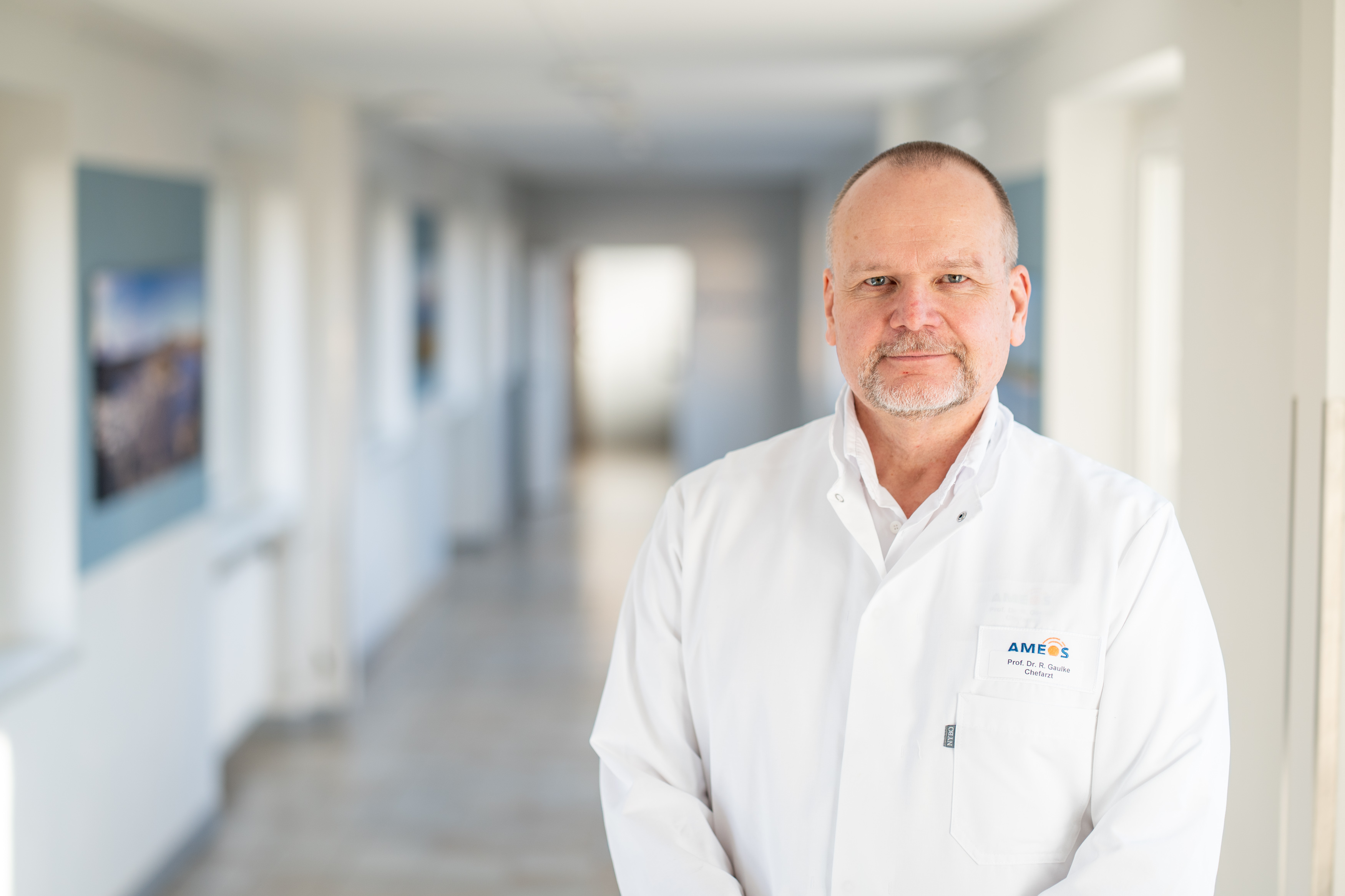 Arzt im Kittel steht in einem Flur und lächelt in die Kamera