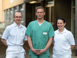 Der neue Chefarzt, der pflegerischer Leiter der Zentralen Notaufnahme und die leitende Oberärztin stehen in Arbeitskleidung vor dem Eingang der Zentralen Notaufnahme am AMEOS Klinikum Halberstadt.